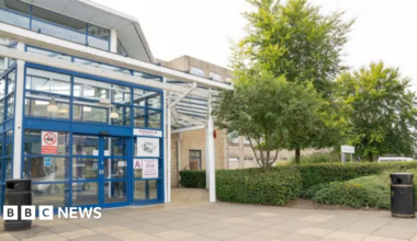 A young man with dark curly brown hair standing by the side of a road. He is wearing a black t-shirt and behind him are yellow crops and blue skies.