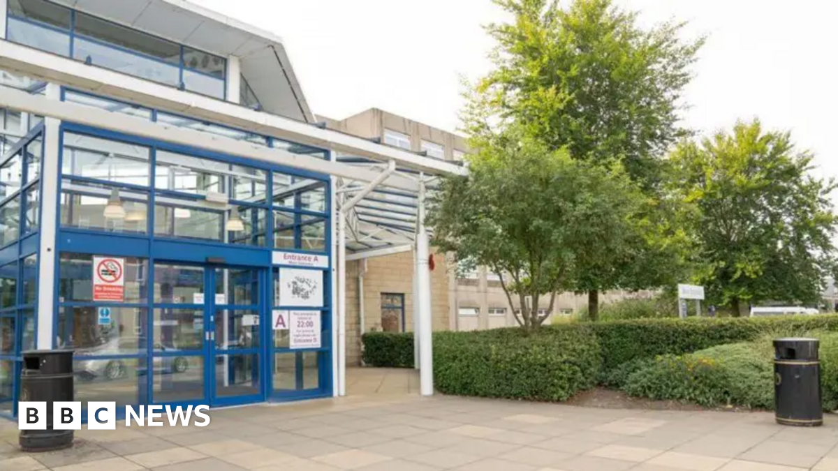 A young man with dark curly brown hair standing by the side of a road. He is wearing a black t-shirt and behind him are yellow crops and blue skies.