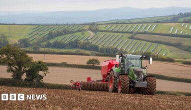 A green and red tractor is pulling a cultivator and drill across a muddy field. On the hills in the background lie solar panels. A light mist sits in the sky.