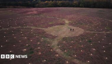A drone image of two people dressed in dark clothing, who are seen from very far away in a clearing. Around them is a large swirling artwork, lit by hundreds of lanterns spreading out in every direction. There are curved paths running through the installation.