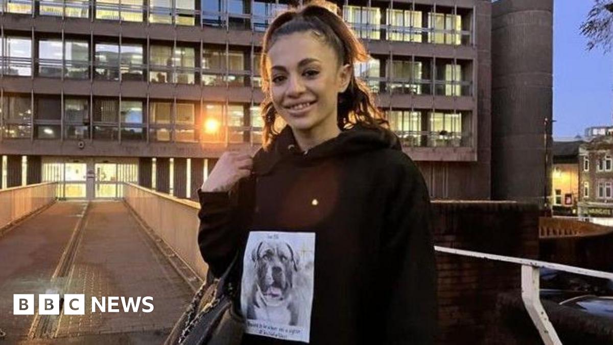 A woman stands outside a grey, brutalist court building at dusk. She is smiling. She has her hair in a high ponytail and holds a handbag at her elbow. She is wearing a black hoodie with a black and white picture of a bulldog on the front.