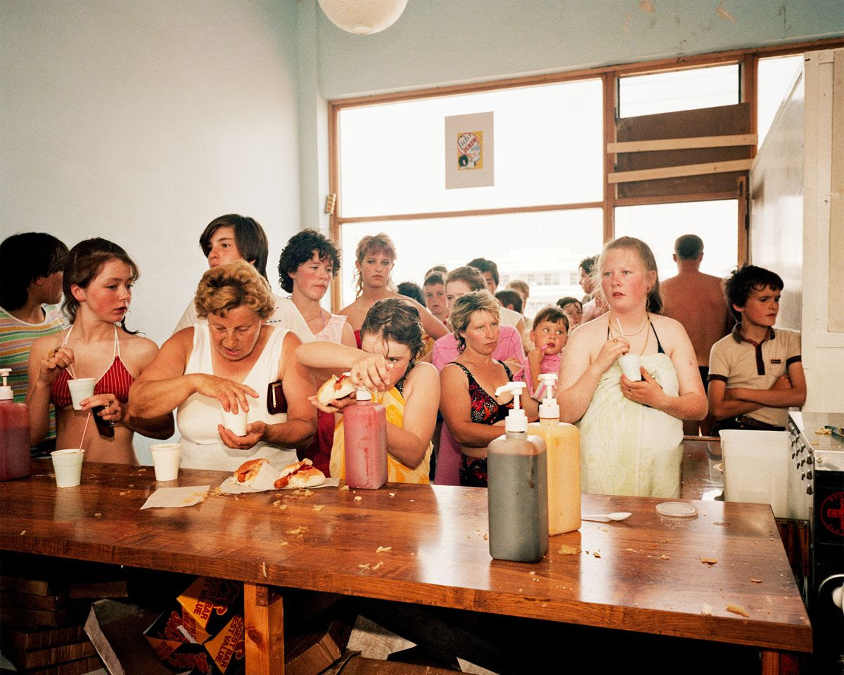 A group of people stands behind a wooden counter in a snack bar, with individuals focused on preparing food and adding condiments from large plastic bottles.