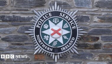 Black, green, and grey circular crest on a grey brick wall, featuring the words Police Service Northern Ireland in white lettering.