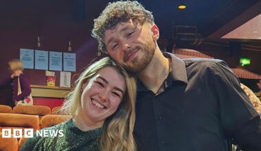Jacob and Lucy inside the cinema. It is a head and shoulders shot. Lucy is on the left and smiles, her long blonde hair is swept to the left and she has a sparkly green top on. Jacob's head is on top of Lucy's, he also smiles and has a black shirt on. There are yellow velvet seats behind as well as a couple of people milling around behind them.
