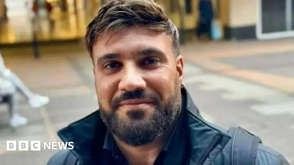 Close up head and shoulders shot of Alastair Chambers wearing a black coat.  He has a beard and a moustache. He is standing in a pedestrianised shopping area.