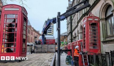 Red telephone boxes are removed in Preston with a large blue crane by a workman in a high viz tangerine jacket and white hard hat.