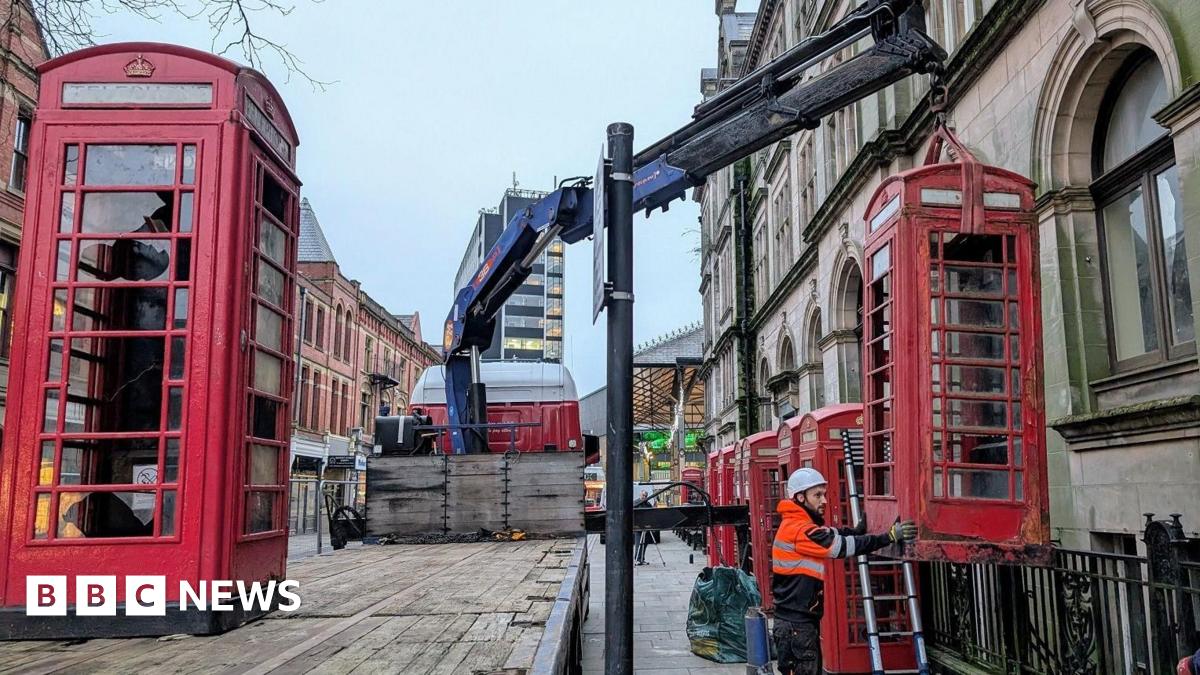 Red telephone boxes are removed in Preston with a large blue crane by a workman in a high viz tangerine jacket and white hard hat.