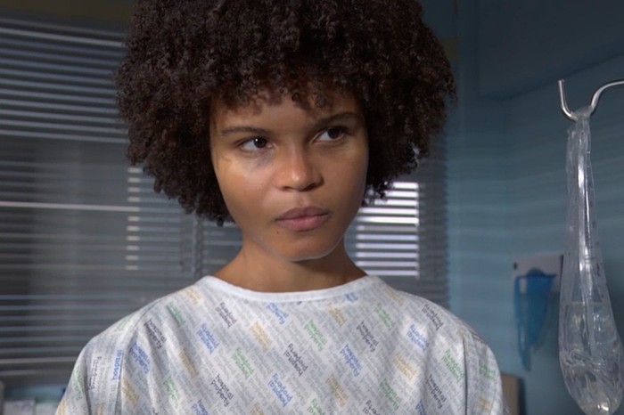Close-up of Indeyarna Donaldson-Holness as Jasmine Fisher in a hospital gown looking pensive, with a window with blinds behind her.