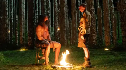 Sony Pictures Ralph Fiennes and another character in a wood either side of small fire