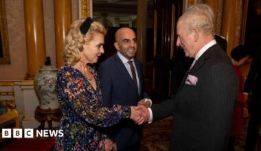 King Charles, wearing a black suit, shakes Eleni Sakkoulei's hand. She has blonde, curled hair and is wearing a flowered dress. Her husband Samir Zitouni stands beside her smiling. An ornate chair, urn, columns, a door, and part of a painting in Buckingham Palace are seen in the background.