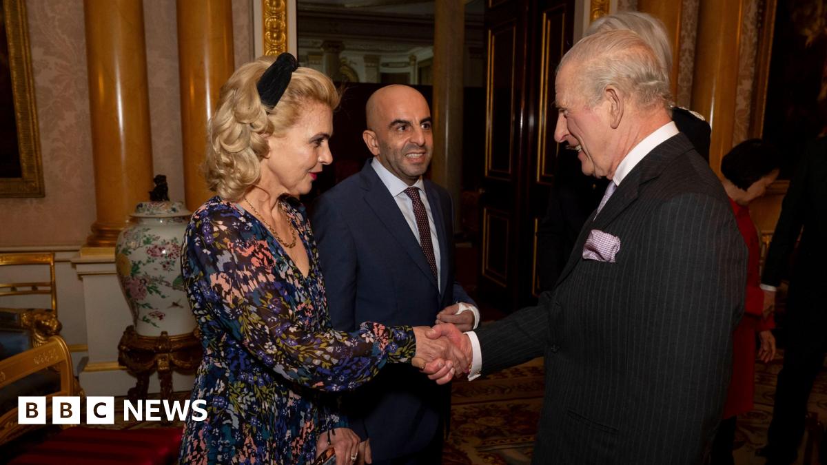 King Charles, wearing a black suit, shakes Eleni Sakkoulei's hand. She has blonde, curled hair and is wearing a flowered dress. Her husband Samir Zitouni stands beside her smiling. An ornate chair, urn, columns, a door, and part of a painting in Buckingham Palace are seen in the background.
