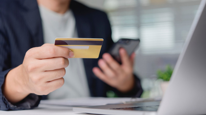 Customer verifies online purchase with credit card and smartphone at office desk near laptop.