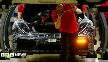 A man in a red tshirt uses a tool on the underside of a car, while a car in the process of being built sits behind him in a factory in Germany.