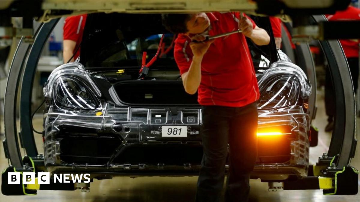 A man in a red tshirt uses a tool on the underside of a car, while a car in the process of being built sits behind him in a factory in Germany.