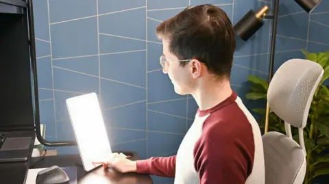 Getty Images A stock image of a young person turning on a Sad lamp on their desk. He is sitting in an office chair and is turned away from the camera, with one finger on the lamp. He is wearing a white and red raglan shirt. He is sitting in front of blue patterned wallpaper.