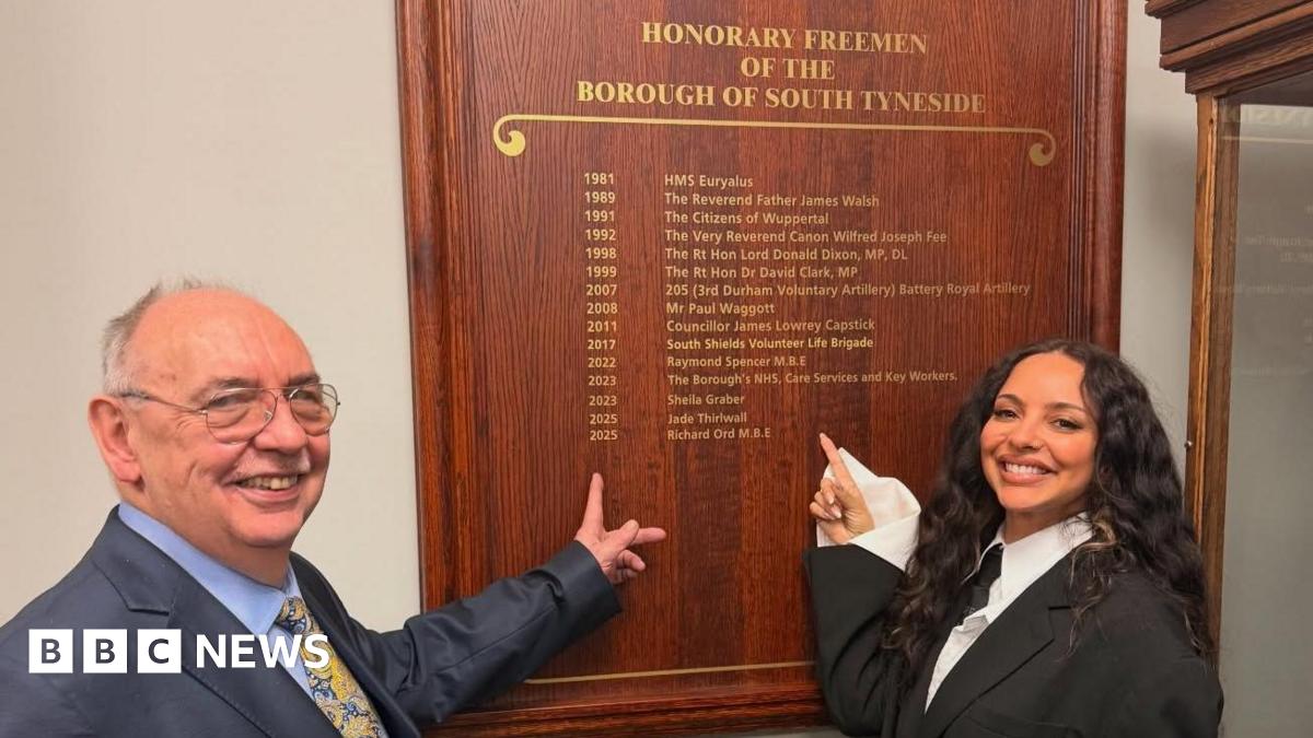 Richard Ord and Jade Thirlwall stand in front of a wooden plaque reading 'Honorary Freemen of the borough of South Tyneside'. Their names are inscribed on the plaque. The pair are pointing at the plaque and smiling.