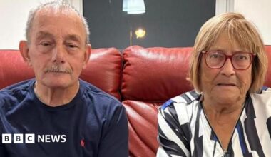 An elderly couple sit on a red leather sofa in a living room. The man on the left has cropped grey hair and a grey moustache, and wears a navy blue t-shirt and dark jeans. The woman has blonde bobbed hair, and wears red-rimmed glasses and a patterned blouse.