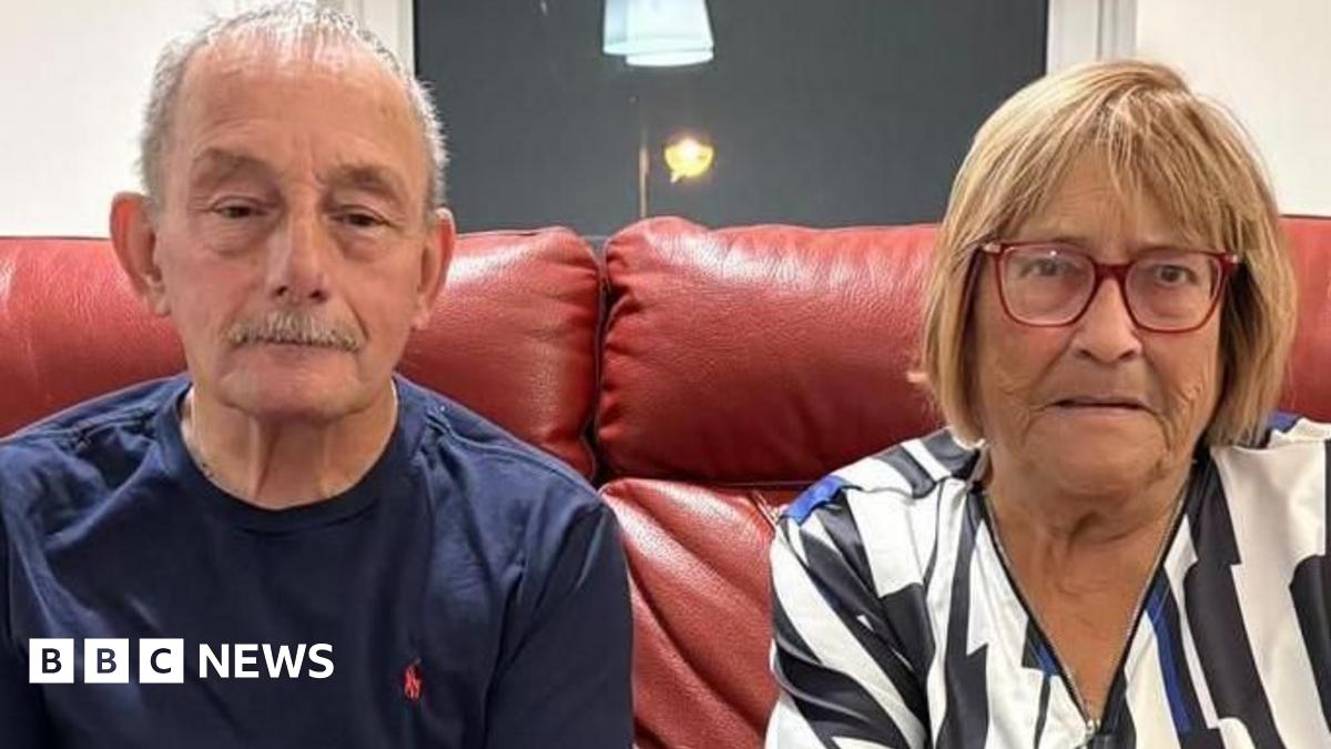 An elderly couple sit on a red leather sofa in a living room. The man on the left has cropped grey hair and a grey moustache, and wears a navy blue t-shirt and dark jeans. The woman has blonde bobbed hair, and wears red-rimmed glasses and a patterned blouse.