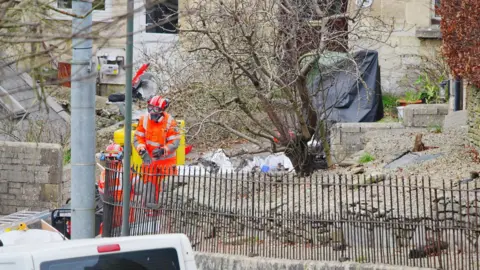 PA Workers dressed in hi-vis orange overalls with masks, goggles and helmets. They are standing in the front yard of the property behind a black iron fence. 