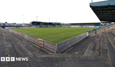 A brown NC500 sign post which says it is 27 miles to Ullapool next to a country road with hills in the background.