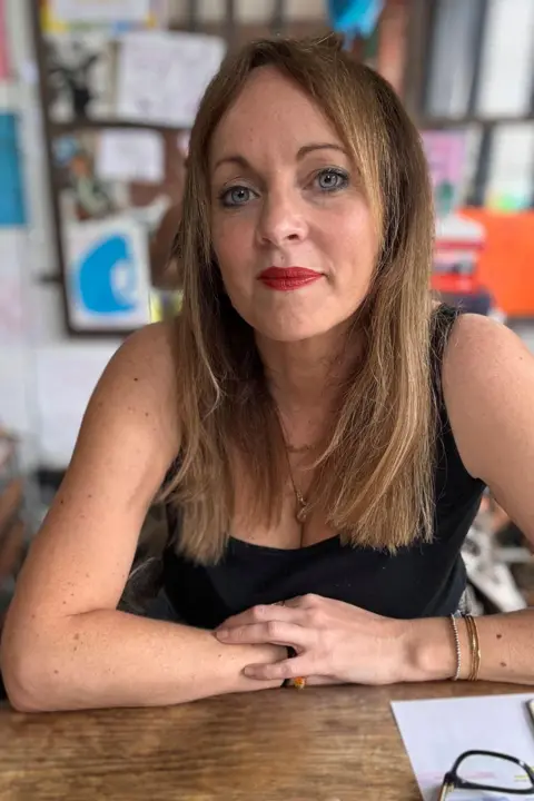 Jenny Evans Jenny Evans looks at the camera, she is wearing red lipstick and has straight blonde hair. She is sat at a table resting her crossed arms on the table. 