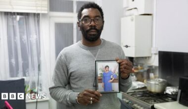 A man in a grey jumper hold photo of his mum in a blue dress. There is kitchen in the background where the hob can be seen.