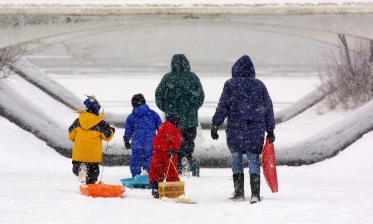 Two adults and three children in winter clothing seen from behind with a toboggan and sliding toys in the snow