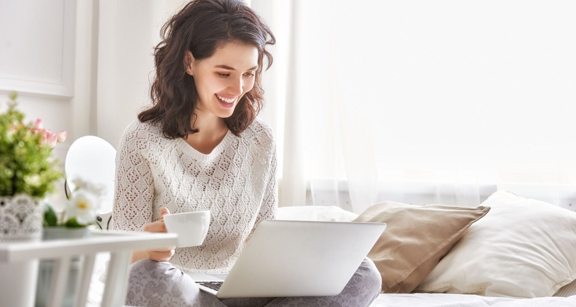 Happy woman sitting on a bed with a coffee and a Windows 11 laptop