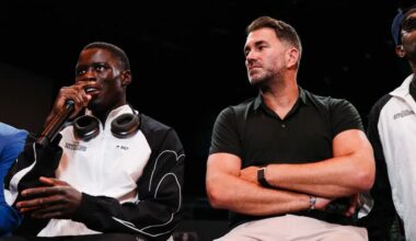 Richardson Hitchins speaks into a microphone during a press event while promoter Eddie Hearn sits beside him with folded arms, listening.
