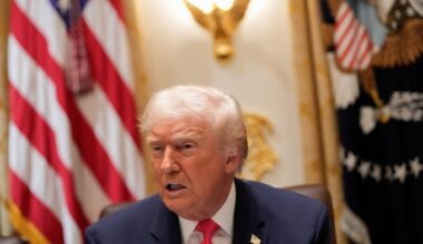 Donald Trump speaks at a table with a serious expression, with the U.S. flag and presidential seal visible in the background.