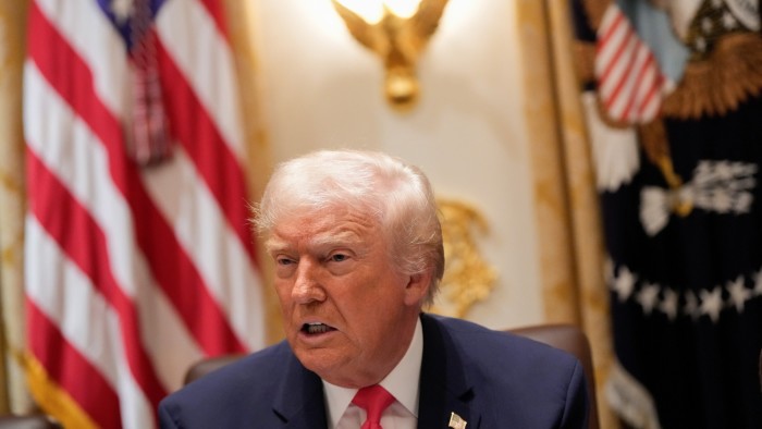 Donald Trump speaks at a table with a serious expression, with the U.S. flag and presidential seal visible in the background.