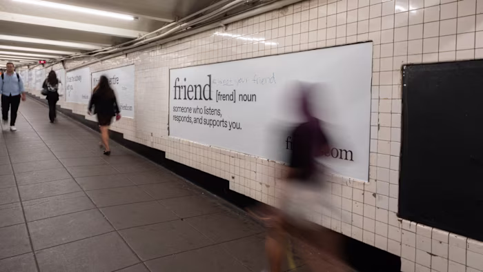 Advertising posters for the “Friend” AI device line a subway corridor, with visible graffiti reading “AI is not your friend.”