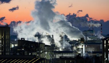 Steam and smoke billow from chimneys at the Korea Zinc Co smelting factory at sunrise, with an orange and blue sky in the background.