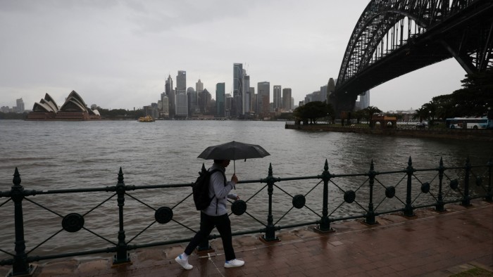 A person with an umbrella walks along a rainy waterfront near the Sydney Harbour Bridge, with the Sydney Opera House and city skyline in the background.