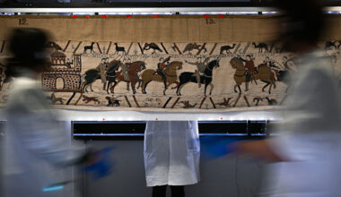 Workers in white coats and gloves stand and move in front of a section of the Bayeux Tapestry, which depicts medieval scenes.