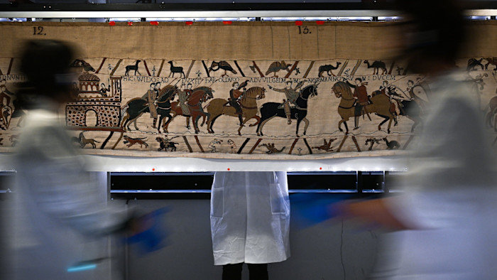 Workers in white coats and gloves stand and move in front of a section of the Bayeux Tapestry, which depicts medieval scenes.