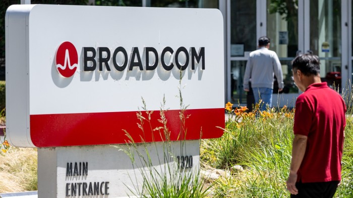 Broadcom headquarters sign with logo and text, with two people walking nearby and office doors in the background.