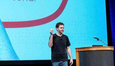 Jonathan Ross stands on stage speaking and gesturing with one hand raised at the GenAI Summit, with a microphone and podium nearby.