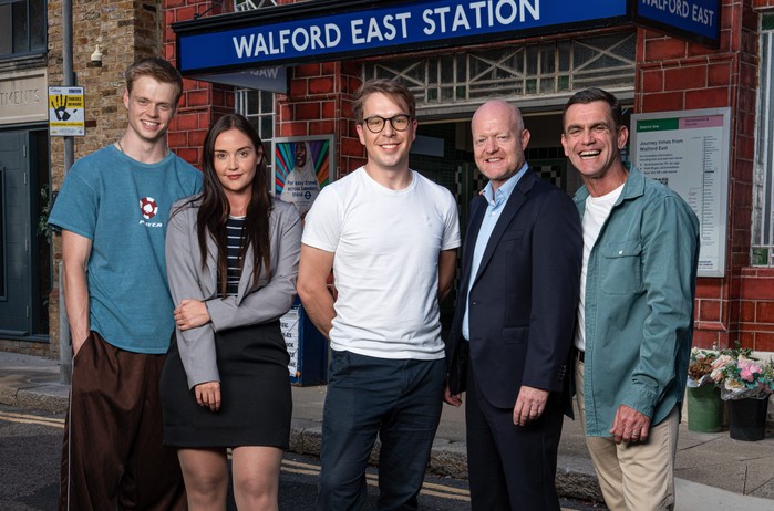 A promo image in front of the Walford East tube station set consisting of smiling, from left to right, Pierre Moullier, Jacqueline Jossa, Ben Wadey, Jake Wood, and Scott Maslen for EastEnders.