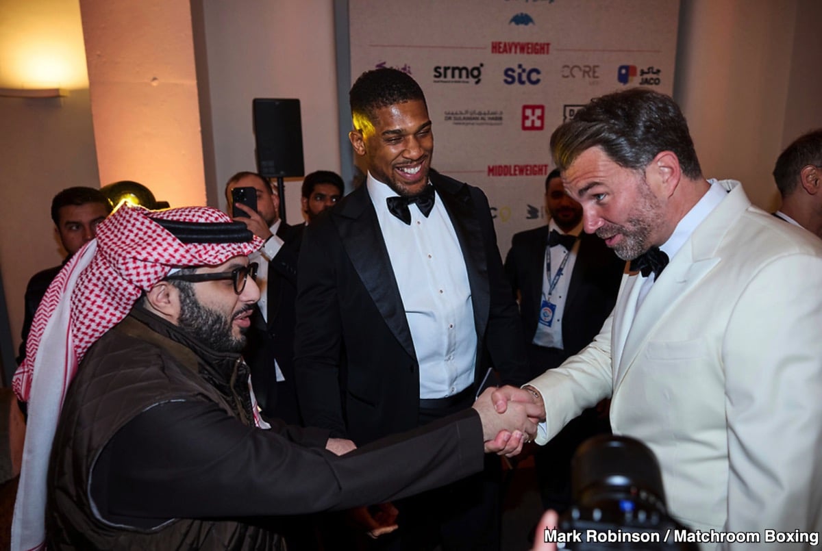 Turki Alalshikh shakes hands with promoter Eddie Hearn during a boxing industry event, as Anthony Joshua looks on in the background.