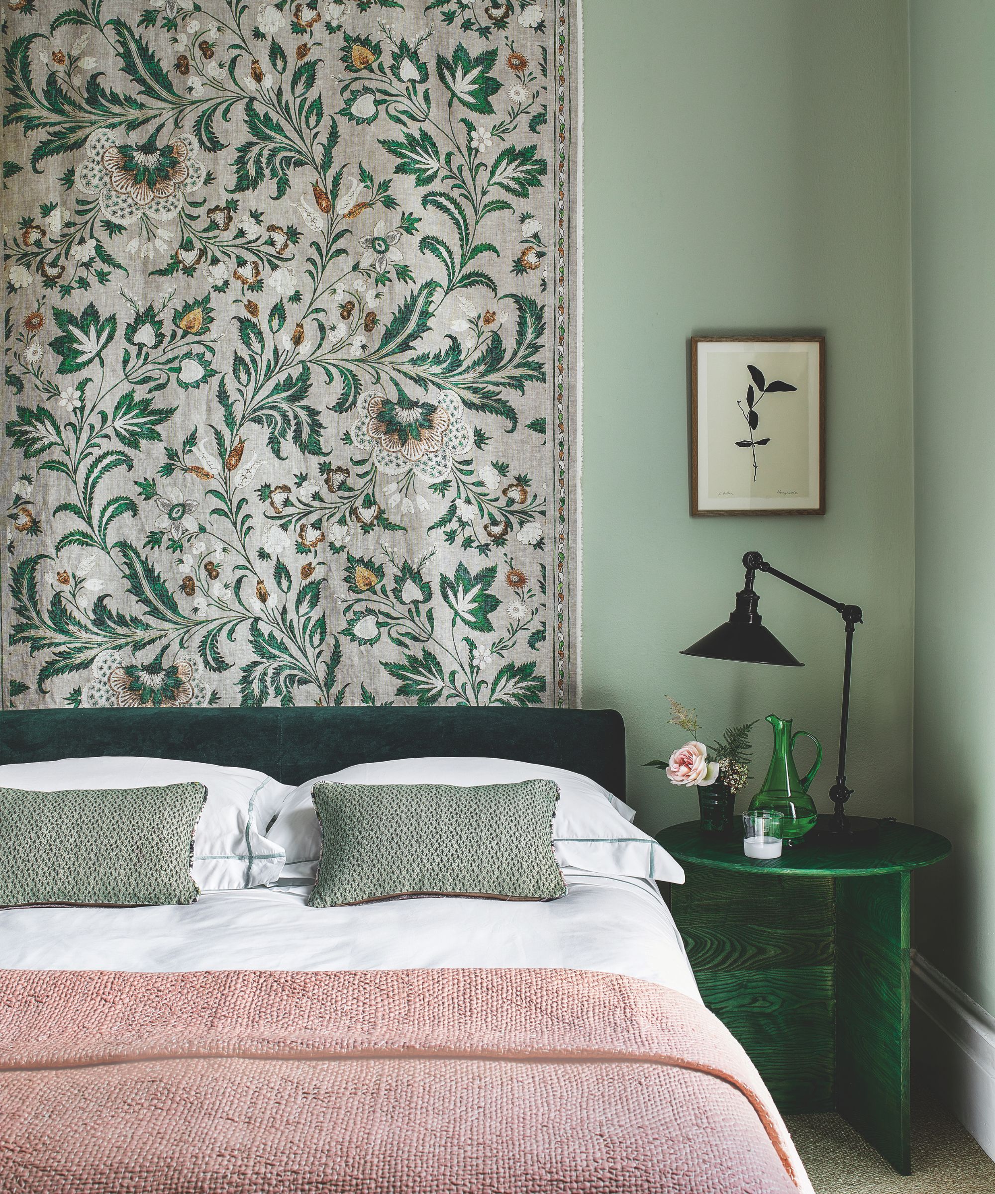 A green bedroom with a green velvet bed with white bedding, a pink blanket and green cushions, a patterned hanging tapestry behind the bed, a green bedside table with a green glass jug, pink flowers and a black lamp, with a small botanical artwork above. The walls are color drenched sage green.