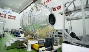 A large metal spacecraft is sits on a platform in a warehouse with lettering behind it