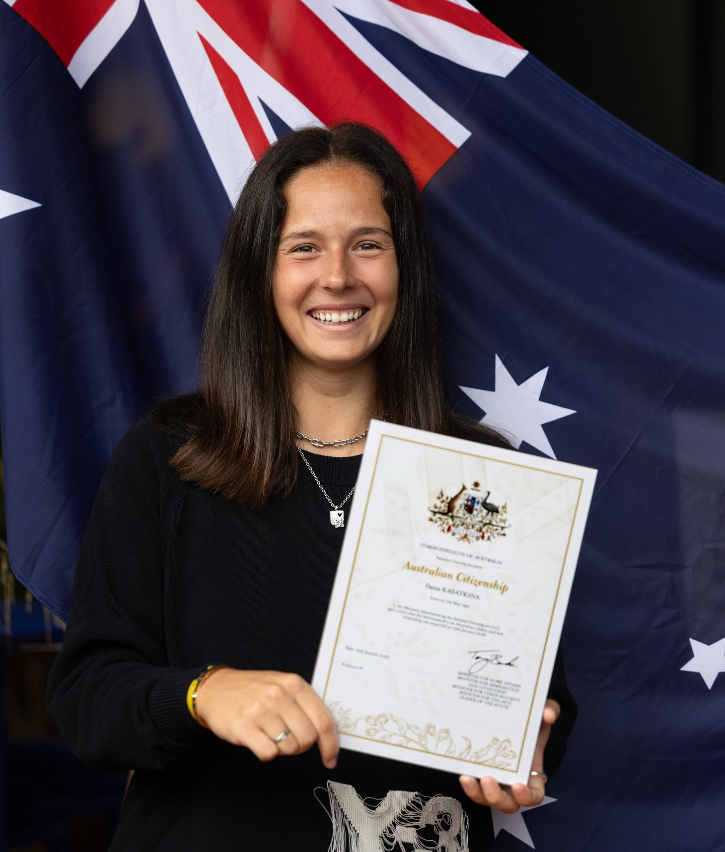 Daria Kasatkina smiling and holding an Australian citizenship certificate in front of the Australian flag.