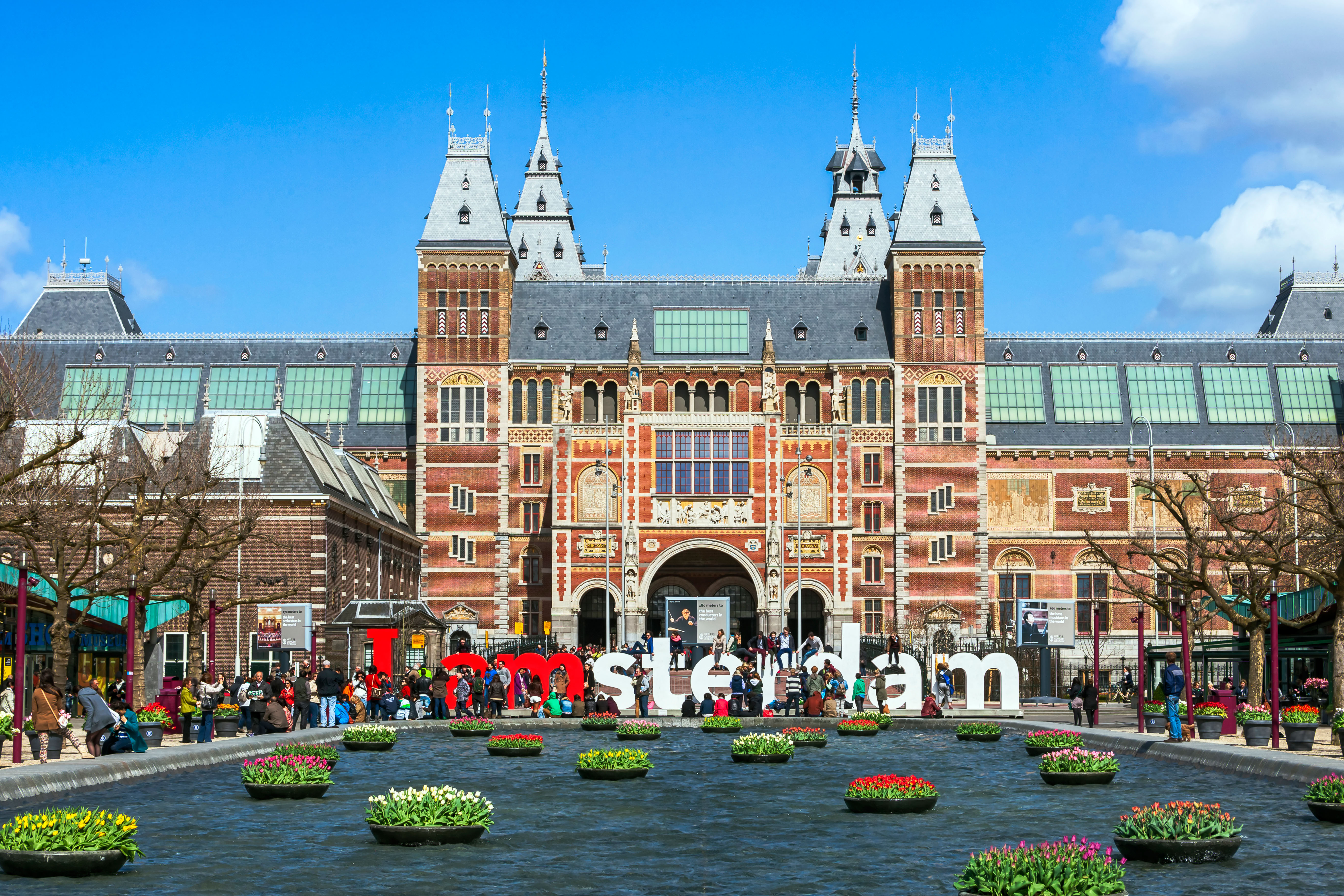 Rijksmuseum in Amsterdam with "I Amsterdam" sign in front, surrounded by tulips and people.
