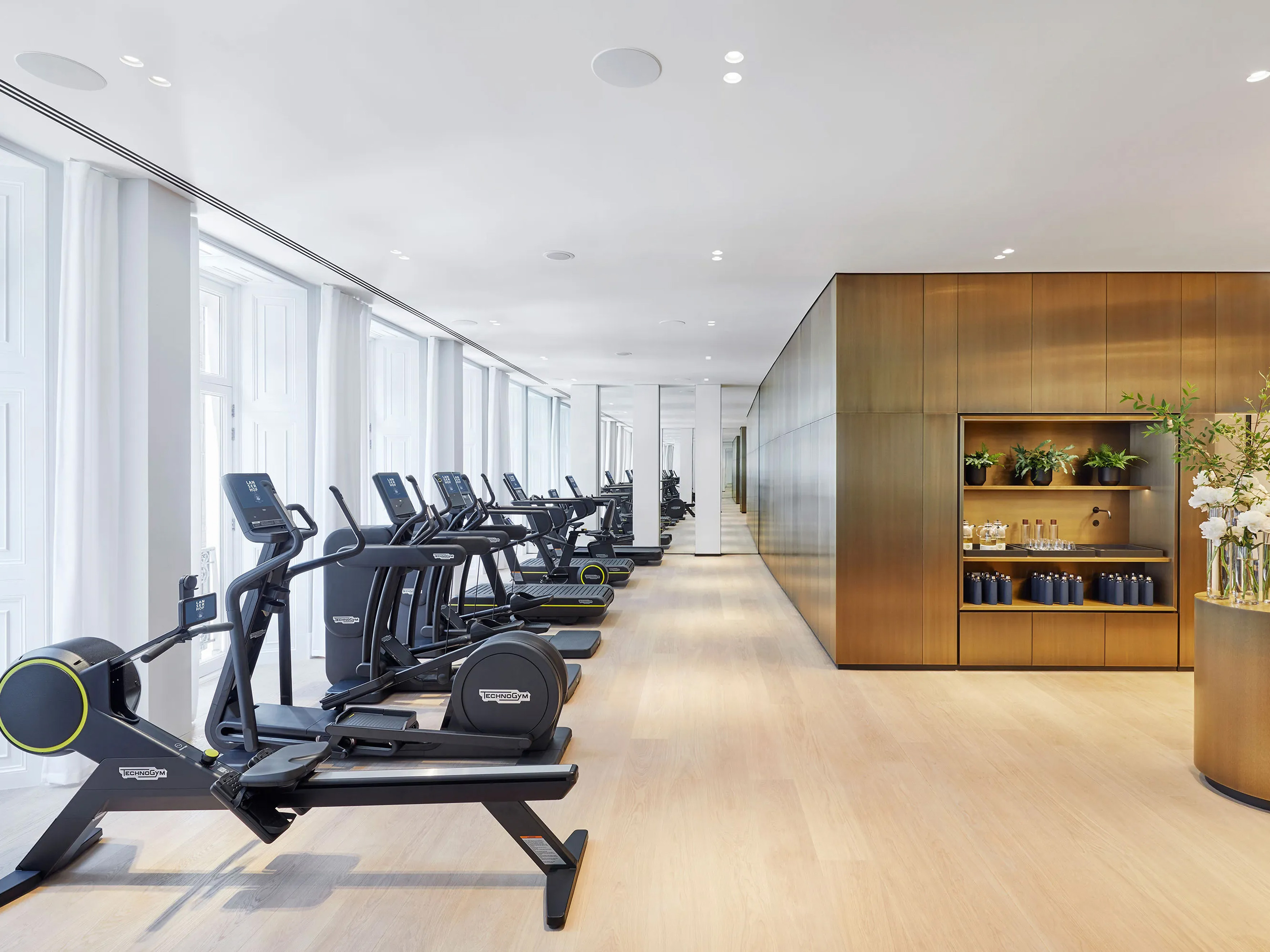 A gym at Lanserhof at The Arts Club with a row of exercise machines facing large windows with white curtains, and a wooden cabinet with plants and bottles on the right.