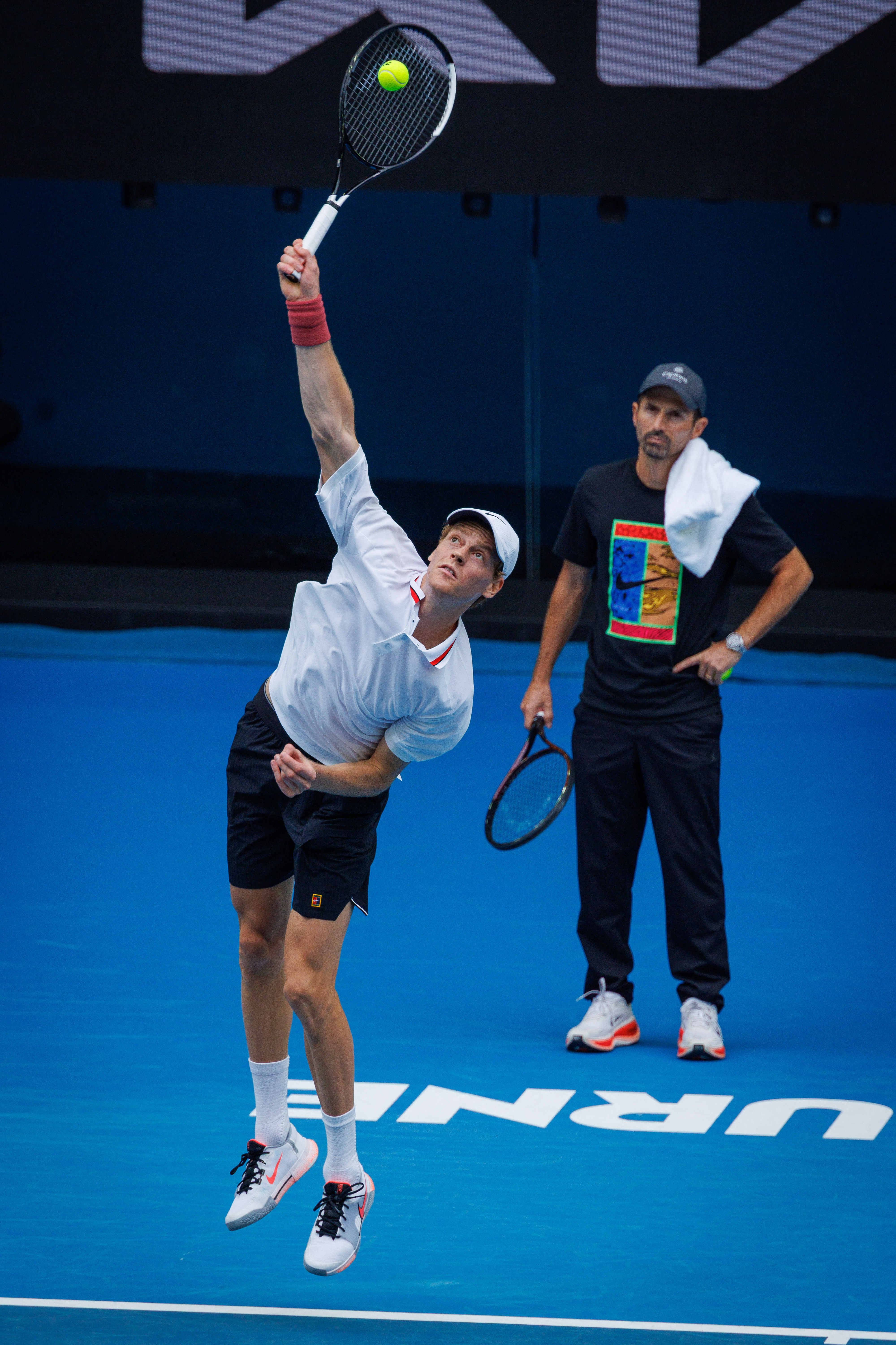 Jannik Sinner serving the tennis ball with his coach Simone Vagnozzi watching.