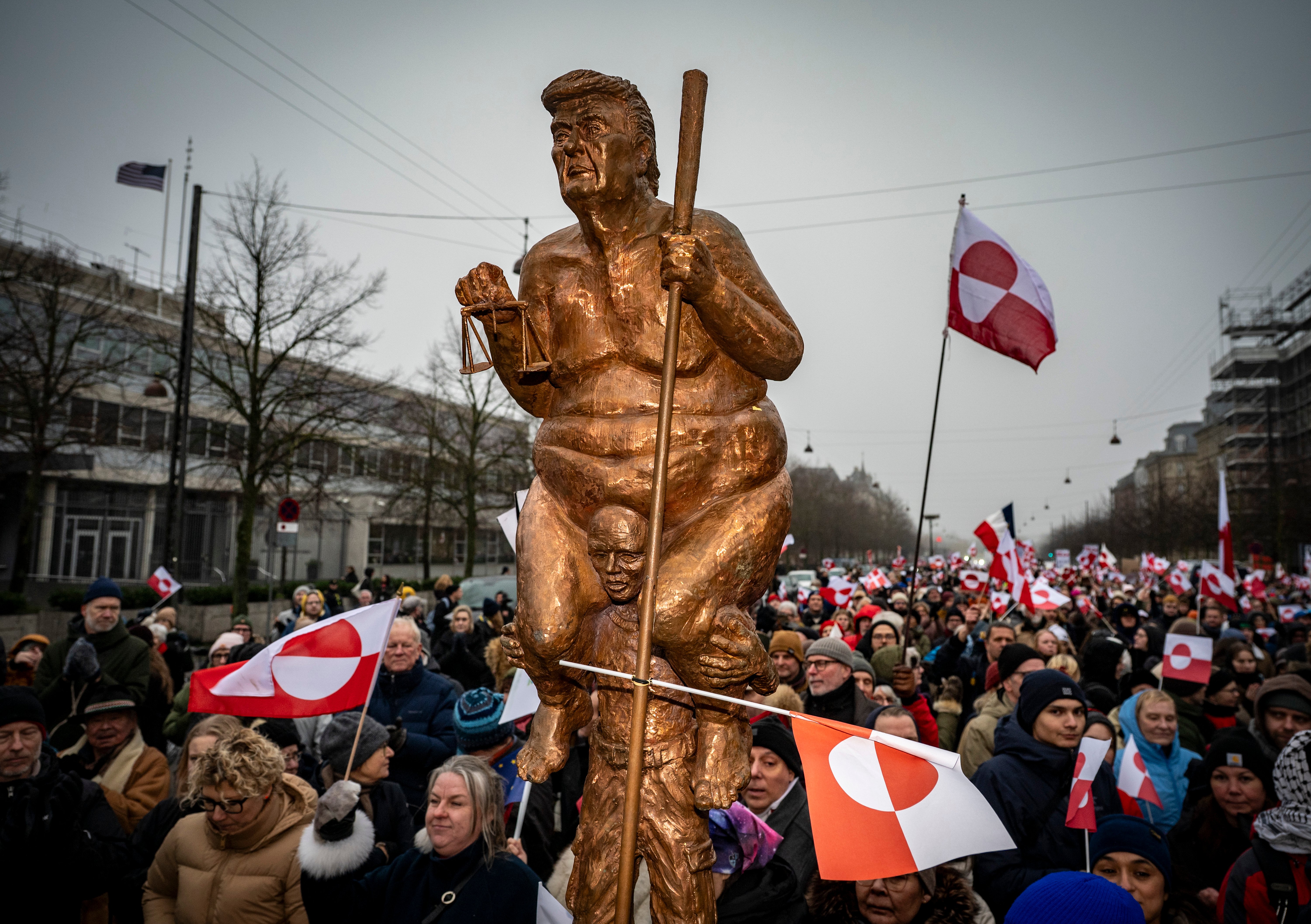 A large bronze statue of Donald Trump holding a scale and a club, carried by a smaller figure, is surrounded by a crowd waving Greenlandic and Danish flags.