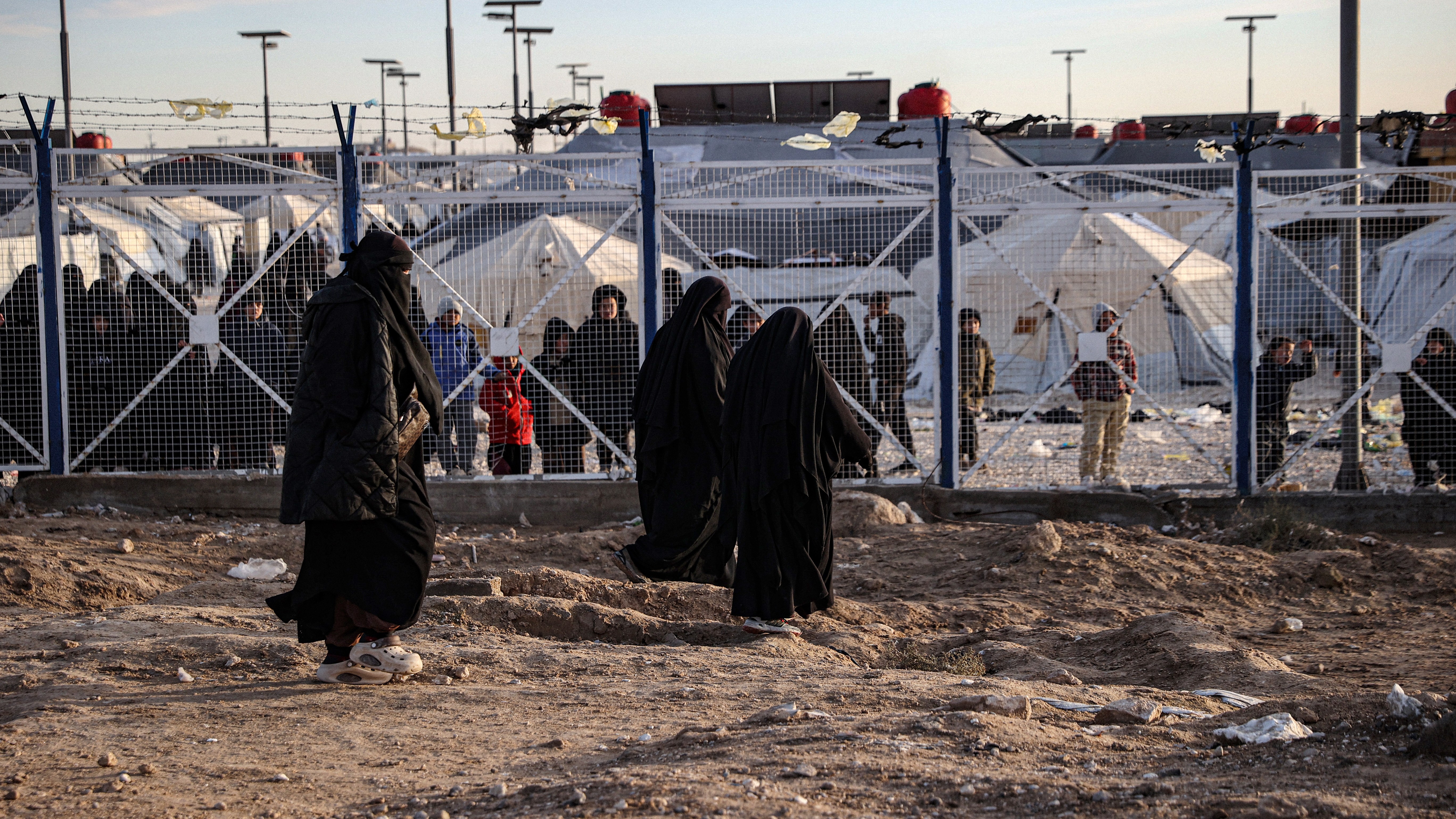 Women in black hijabs walk in front of a fenced camp where tents and children are visible.