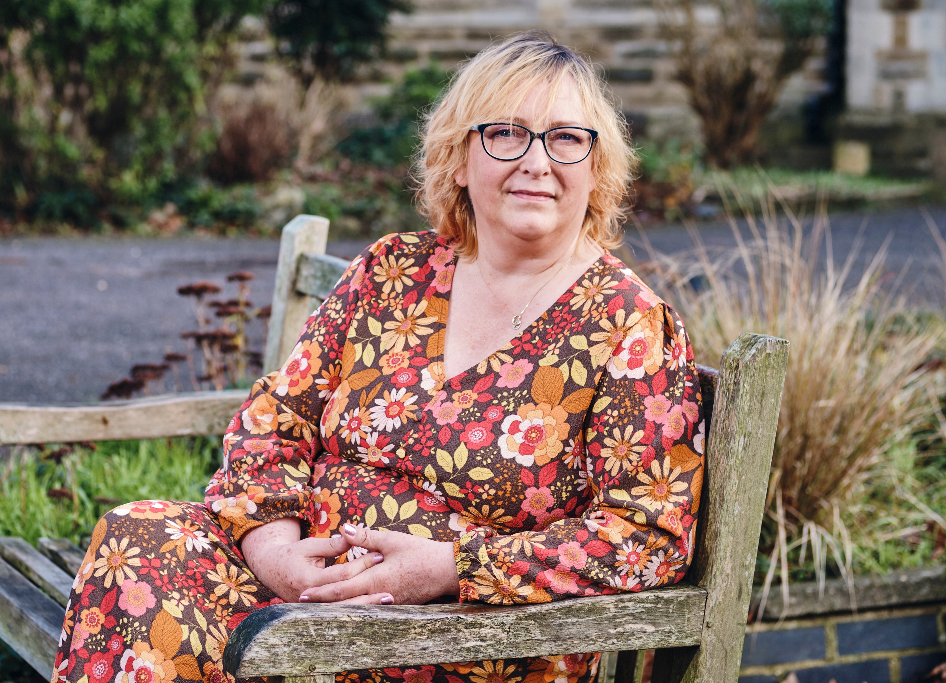 Amy Schmidt sitting on a wooden bench, wearing a floral dress, and glasses.