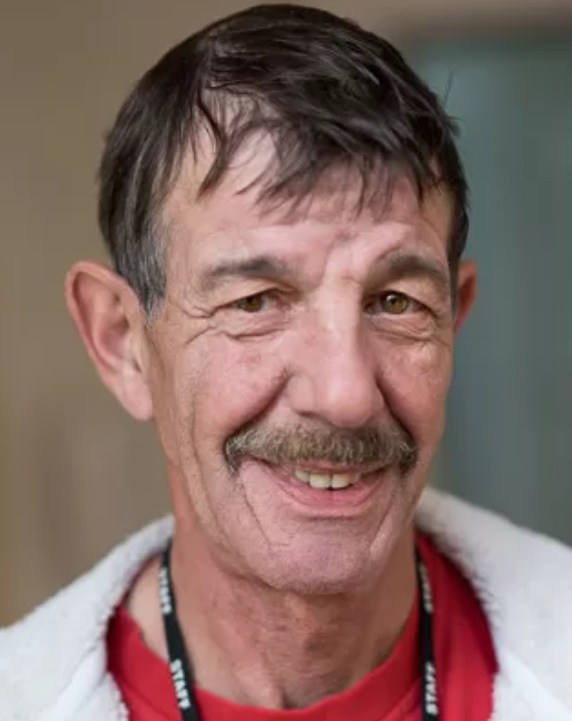 Headshot of Ian Coates, a man with a mustache, smiling and wearing a red shirt and white jacket, with a black "STAFF" lanyard visible.